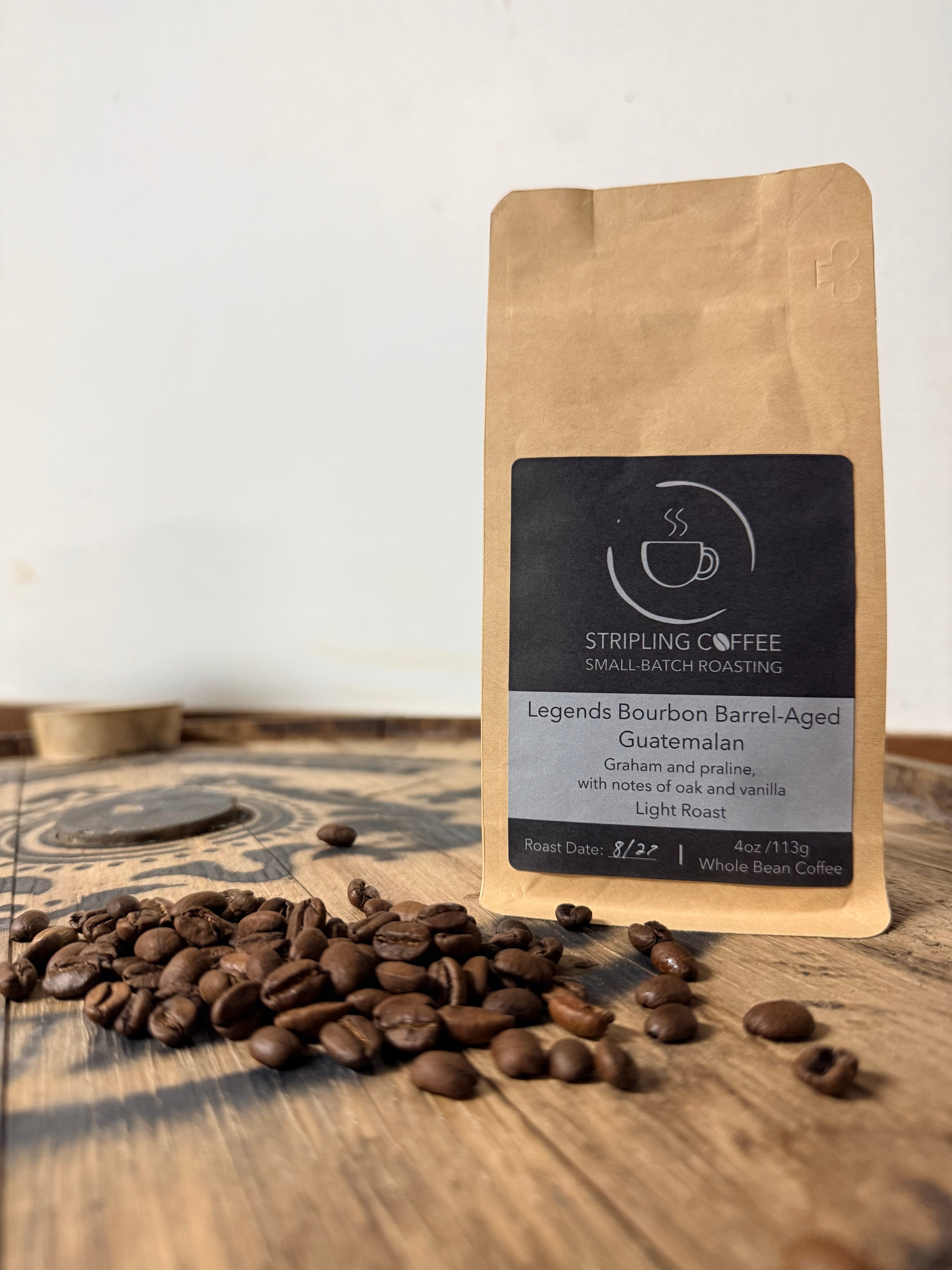 Bag of Stripling Coffee Legends Bourbon Barrel-Aged Guatemalan, light roast, on a wooden table with roasted coffee beans spilled in front of it.